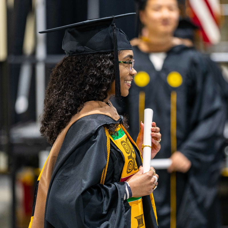 A graduate wearing a cap and gown, holding a diploma scroll and posing to the side